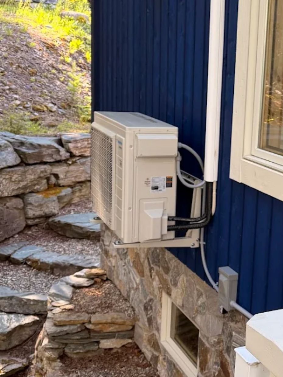 A heat pump's outdoor unit on a wall bracket above the stone foundation of a blue house, on a sunny summer day.