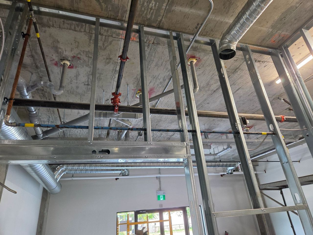 Pipes and ducting running across the concrete ceiling of a commercial building, above new steel stud framing, before the drywall goes up.
