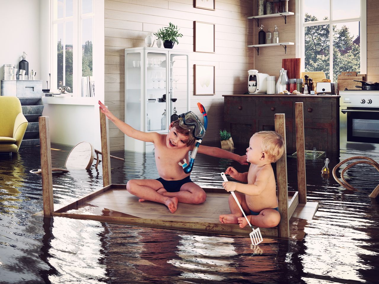 Two kids having fun on an upturned kitchen table floating in a flooded kitchen, the older one in a snorkel mask.