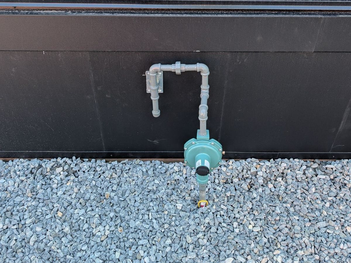 A gas line rising out of the gravel through its regulator and fastened to the foundation wall of a house.