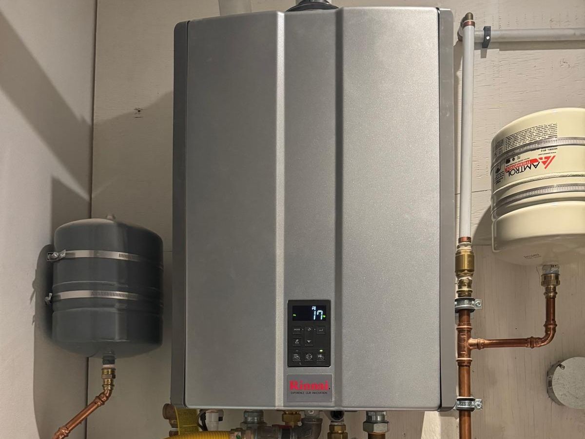 A wall-mounted Rinnai combi boiler that heats both the water and the in-floor heat, with its copper piping, valves, and expansion tanks laid out neatly around it.